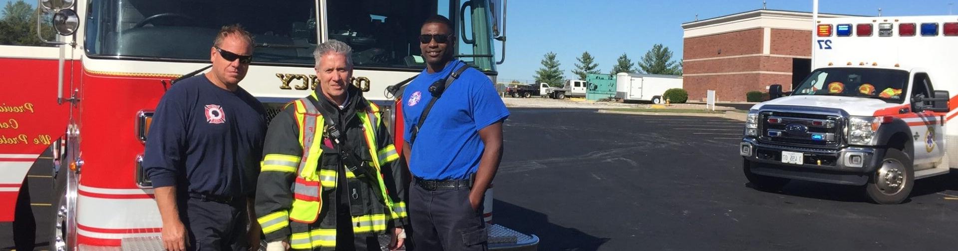 A police officer, firefighter, and medic pose in front of emergency vehicles
